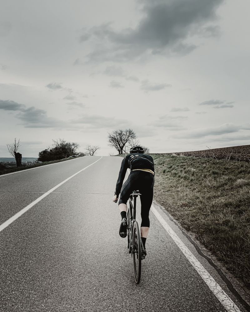 Ciclista de espaldas subiendo por una carretera vacía bajo un cielo gris
